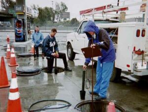 Workers in rain gear inspecting underground storage tanks at a gas station in wet weather.