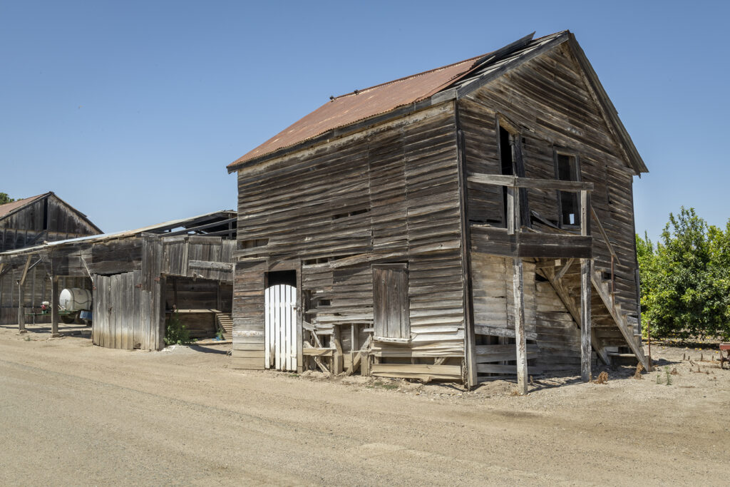 An old, weathered wooden building with a rusted roof, located on a dusty road under a clear blue sky.