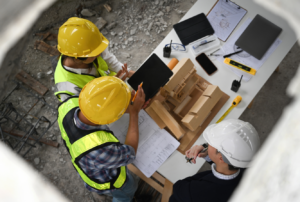 Three construction workers wearing helmets and high-visibility vests examining plans and a model on a table at a construction site.