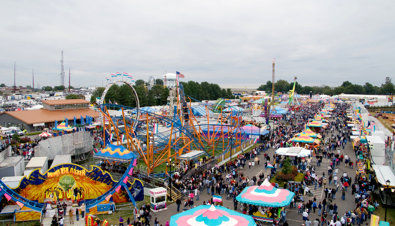 Aerial view of a local fairground with various rides, including a large roller coaster, tents with colorful tops, and a crowd of people walking through the fair.