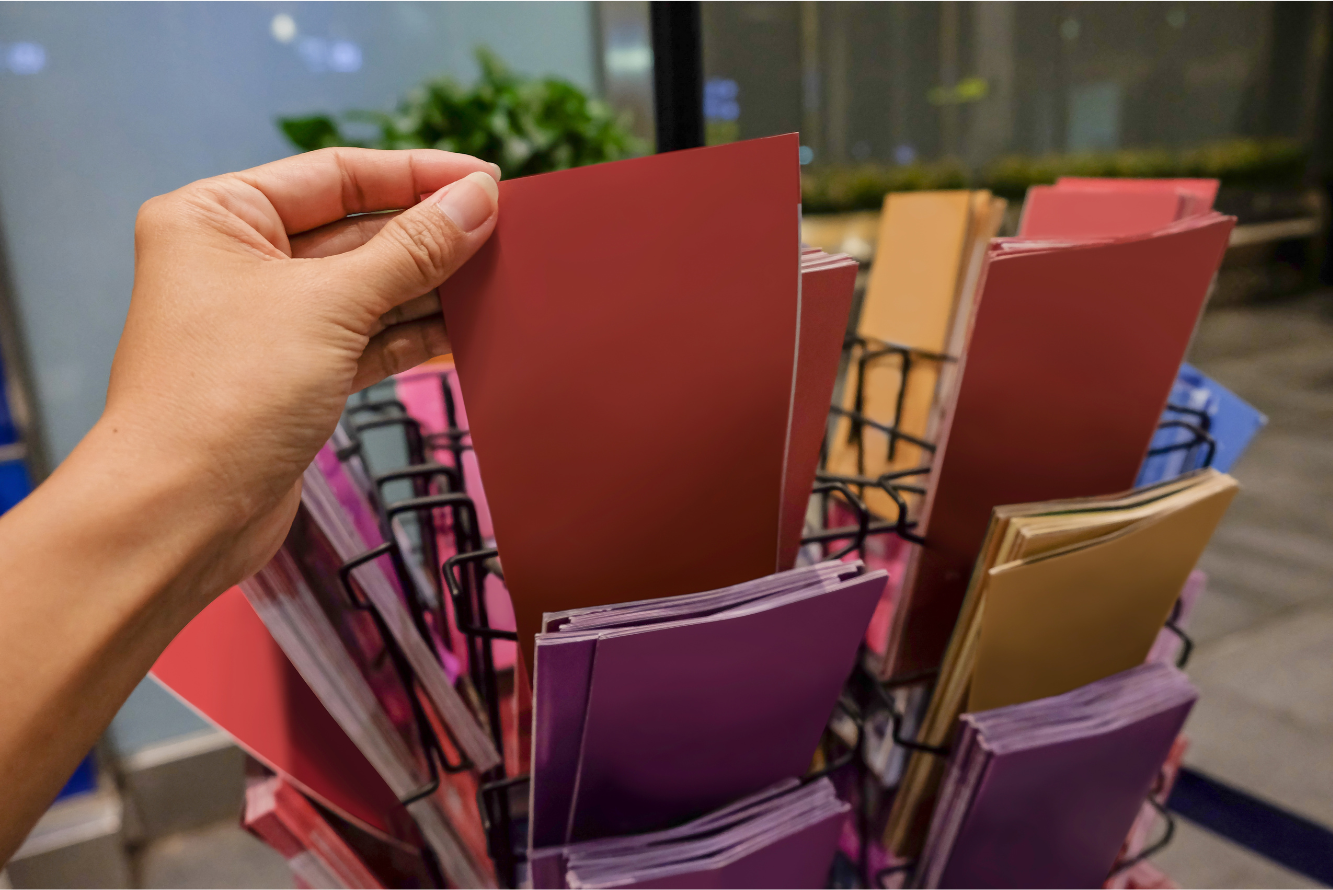 A person’s garbing holding a red-colored brochure in front of a rotary stand filled with brochures of various colors including purple, gold, and blue.