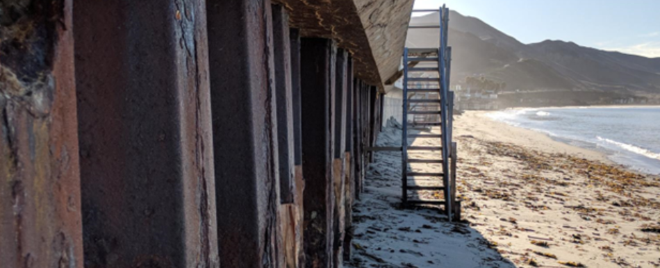 Rusted steel beams and stairs leading to a sandy beach with mountains in the background.