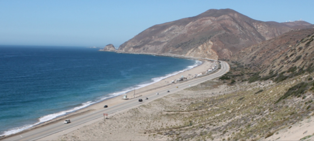 Coastal highway along a scenic beach with mountains in the background.