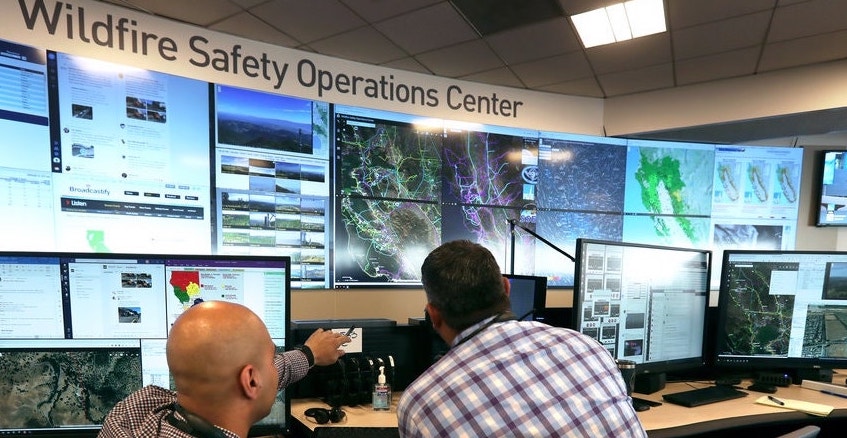 Two men in a wildfire safety operations center observe large monitors displaying weather conditions, maps, and fire data for real-time monitoring and response.