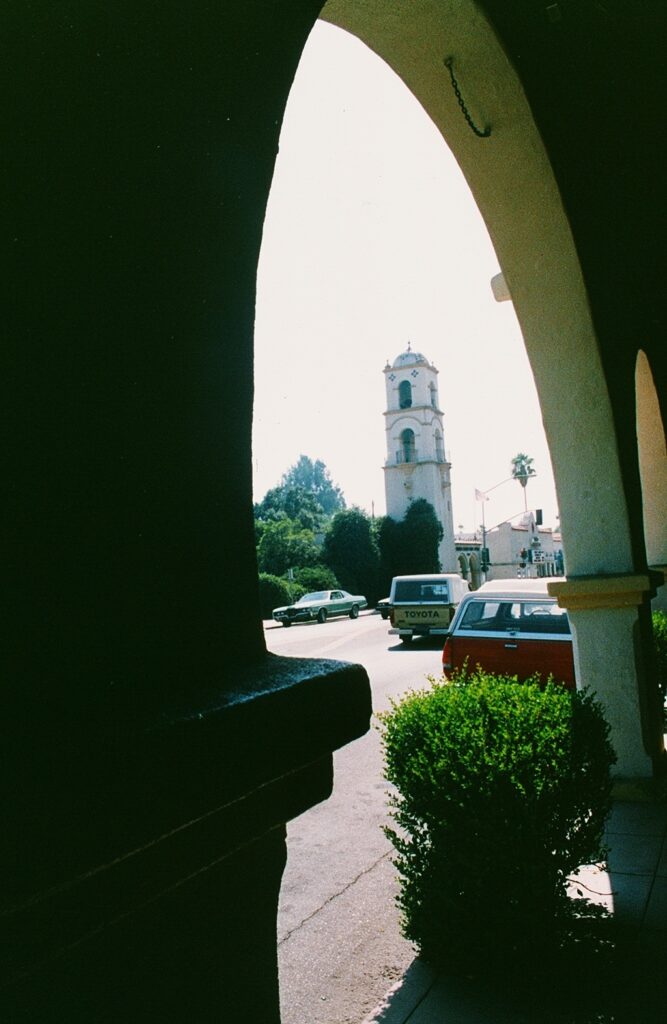 View of Ojai Post Office Tower and Portico in 1985 through an archway, with cars and greenery in the foreground.