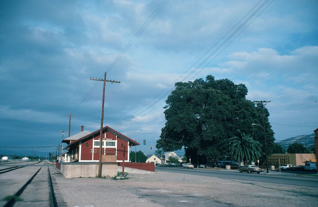 Santa Paula Railroad Depot in 1982, showing the red building beside train tracks, with large trees and a street with cars in the background.