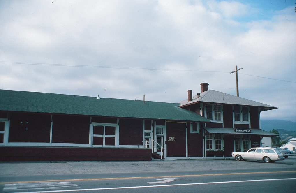 Side view of Santa Paula Railroad Depot in 1982, featuring a long red building with a green roof and a white station wagon parked in front.