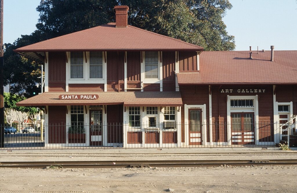 Front view of Santa Paula Railroad Depot in 1985, featuring a red building with "Santa Paula" and "Art Gallery" signs, and train tracks in the foreground.