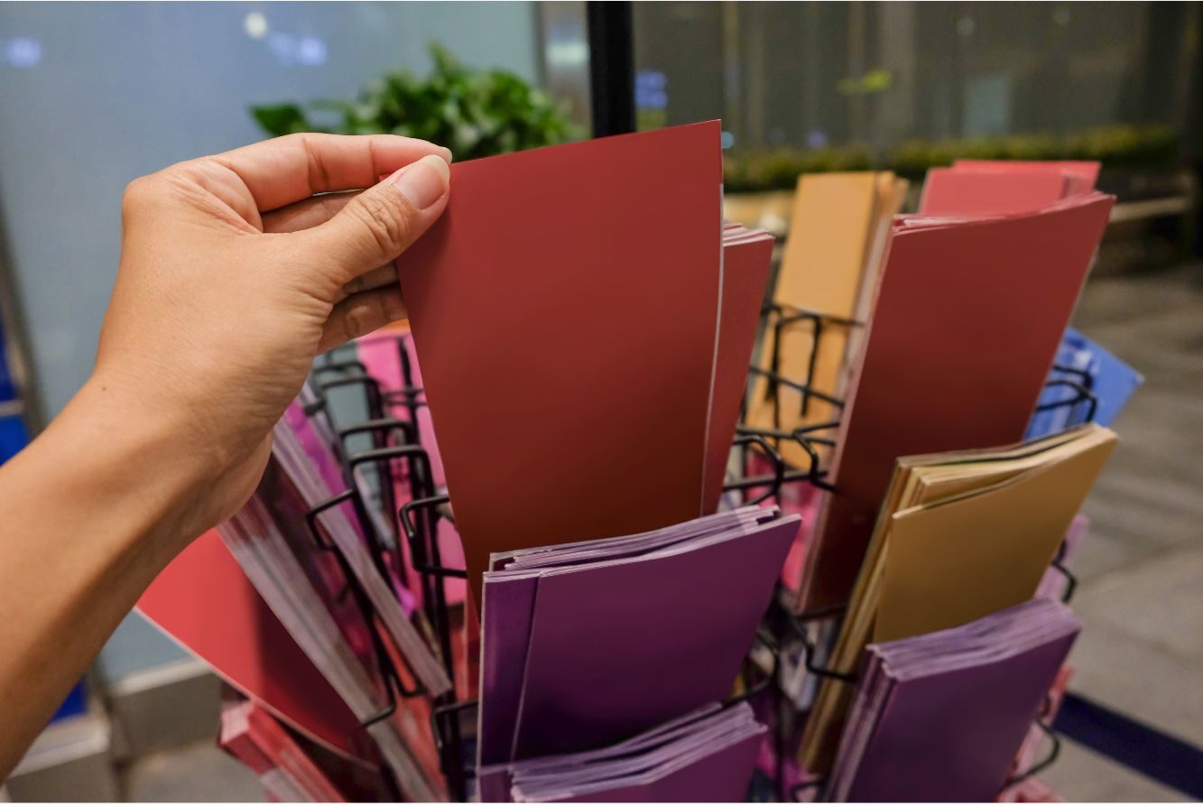 A person’s garbing holding a red-colored brochure in front of a rotary stand filled with brochures of various colors including purple, gold, and blue.