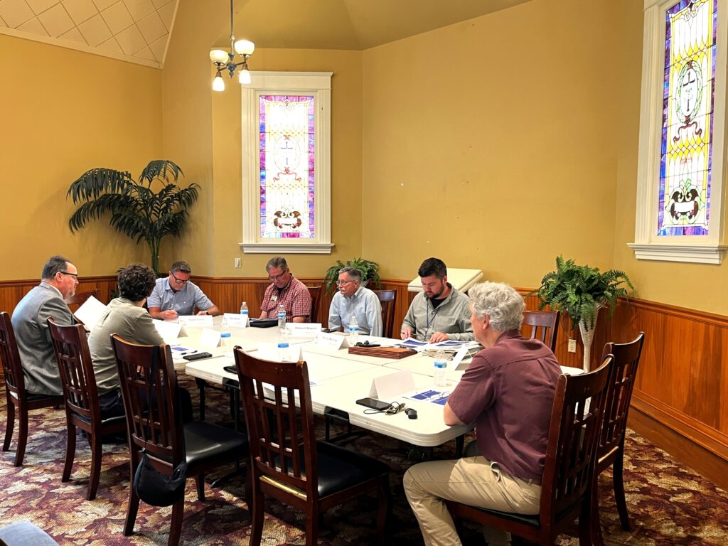 People seated around a table with documents at a meeting in a room with stained glass windows.