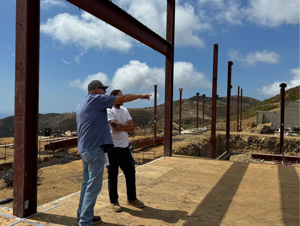 Two men discussing and pointing at a construction site with steel beams around them.