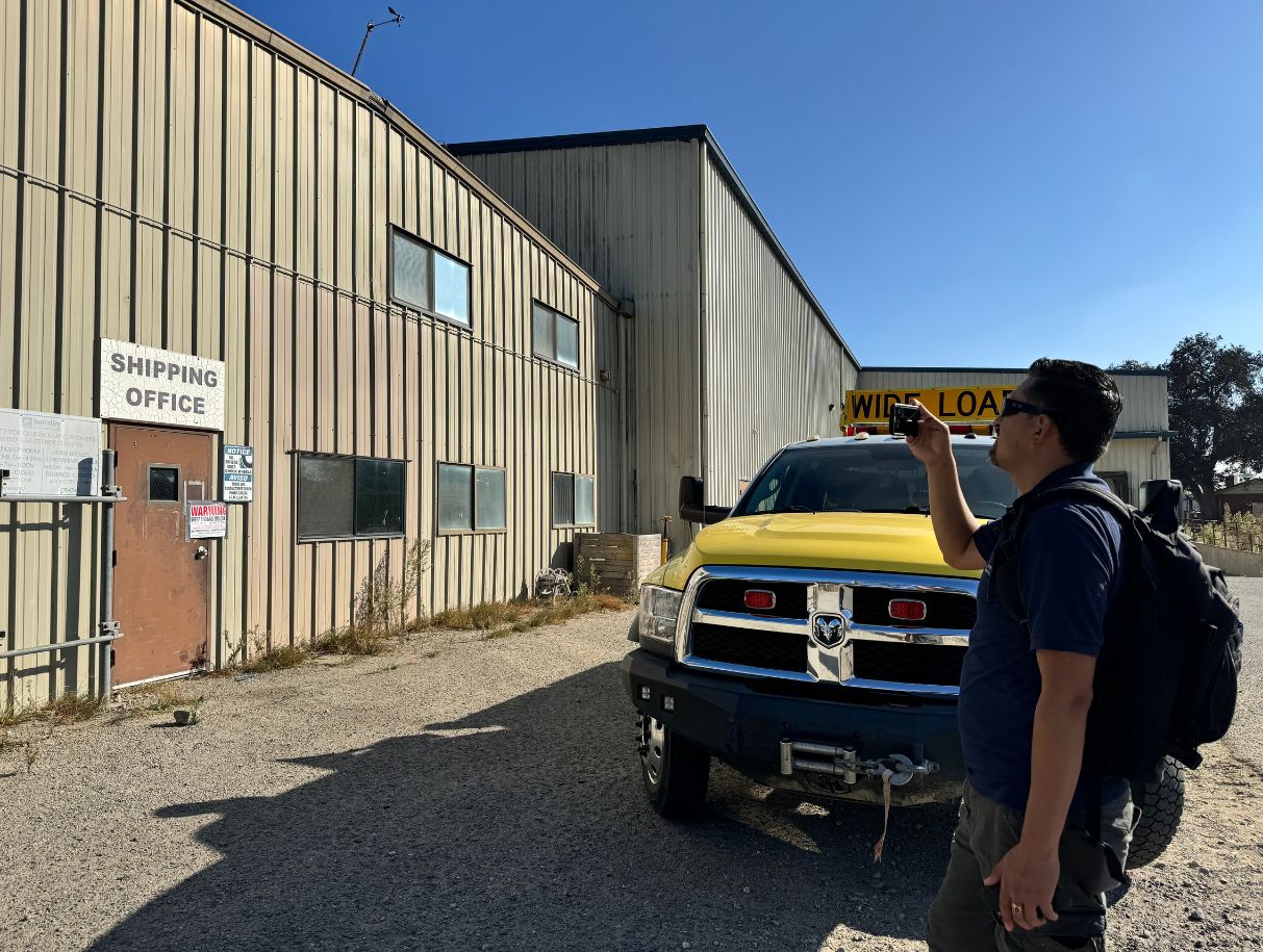 Person with a backpack stands next to a yellow truck taking a picture outside of the shipping office.