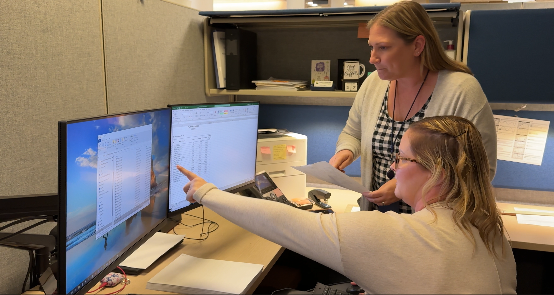 Two office workers reviewing documents and data on dual monitors.