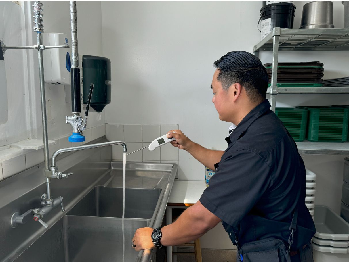 A person in uniform testing water temperature at a stainless steel sink using a digital thermometer.