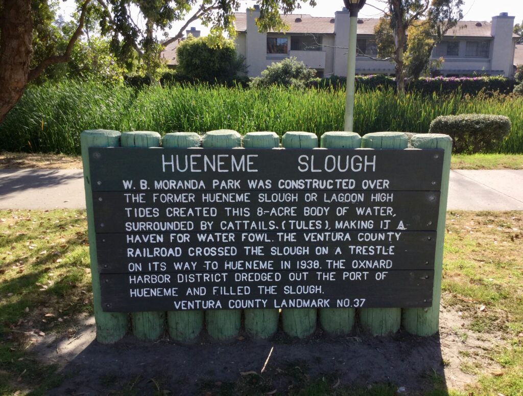 Hueneme Slough Site marker with historical details and greenery in the background.