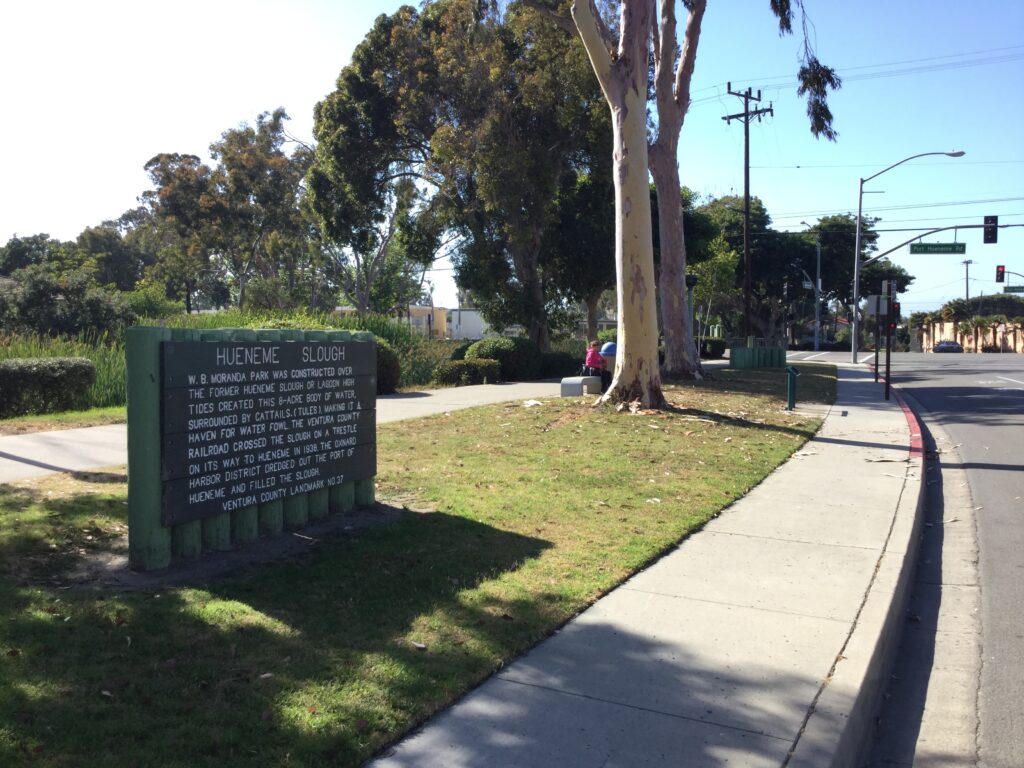Hueneme Slough Site marker with text detailing history, next to a sidewalk and trees.