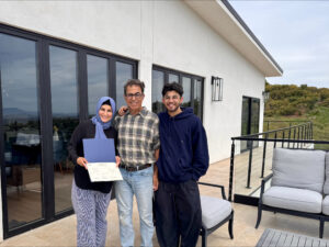 Tres personas sonriendo y posando juntas en el patio de una casa, con una persona sosteniendo un certificado o documento oficial, frente a una moderna casa de estuco blanco con ventanas grandes y una vista panorámica de las colinas al fondo.