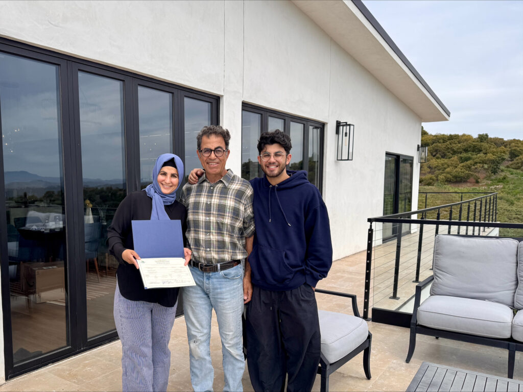 Three people smiling and posing together on a home patio, with one person holding an official certificate or document, set against a modern white stucco home with large windows and a scenic hillside view in the background.