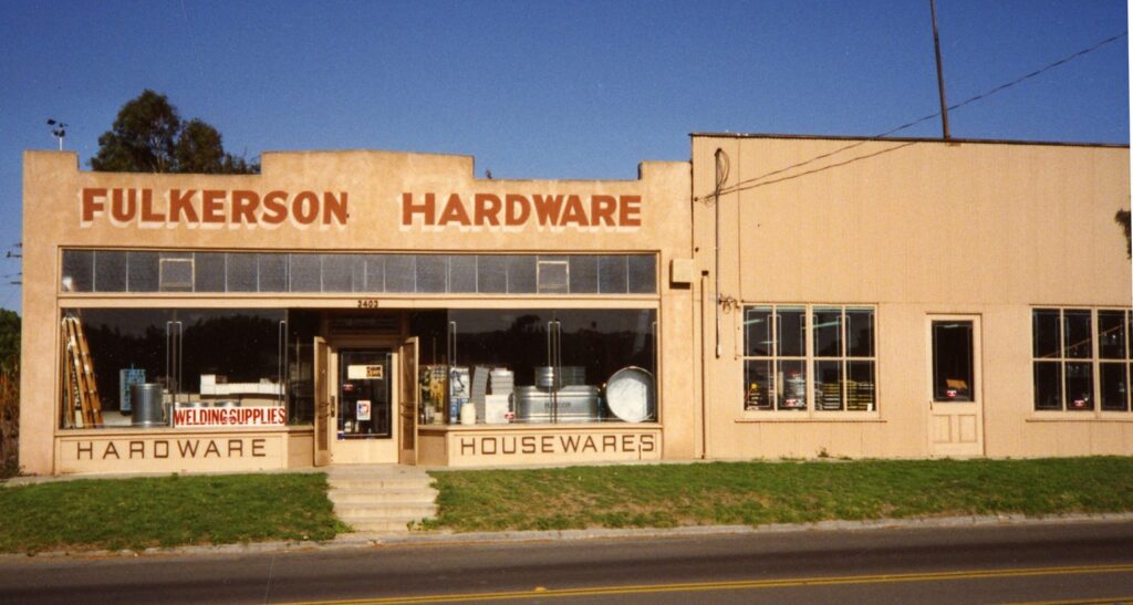 Fulkerson Hardware storefront from the 1980s features red signage for "FULKERSON HARDWARE", "WELDING SUPPLIES", and "HOUSEWARES" on a beige building with large windows showing hardware items.