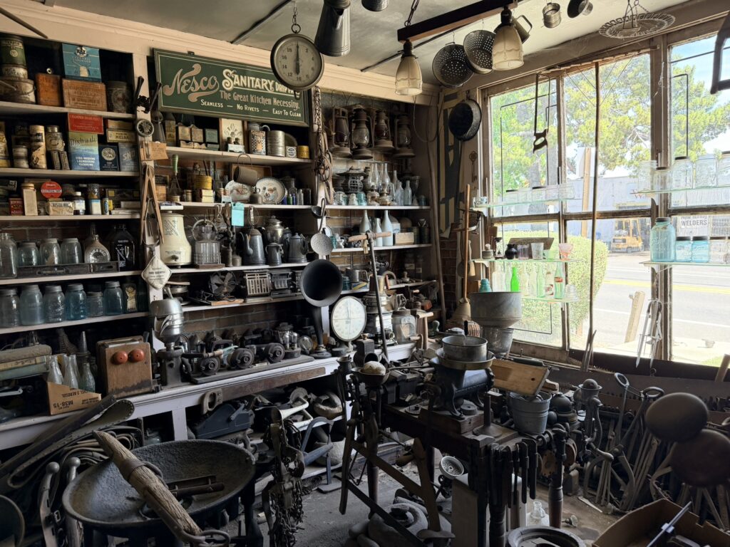 A vintage hardware store museum interior filled with antique tools, kitchenware, and a green "Nesco Sanitary" sign on shelves, viewed through large windows overlooking a street.