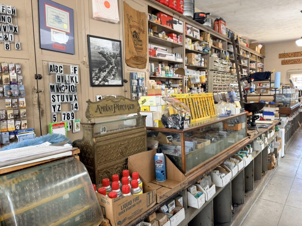 Vintage cash register labeled "Amount Purchased" in a hardware store, surrounded by shelves of goods, letter tiles, and a "BRACKET HARDWARE" sign. Visible items include a box of liquid fire quick starting fluid and glass display cases.