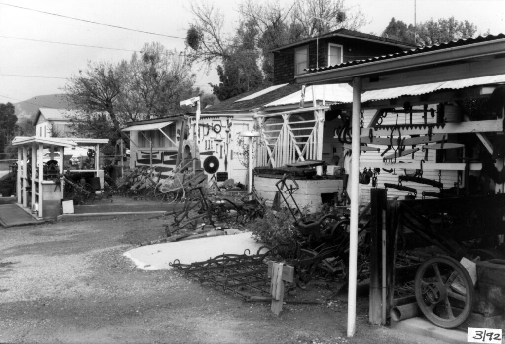 Black-and-white photograph of Lechler Museum's 1992 outdoor exhibit featuring antique agricultural tools, machinery, and hanging implements arranged around buildings.