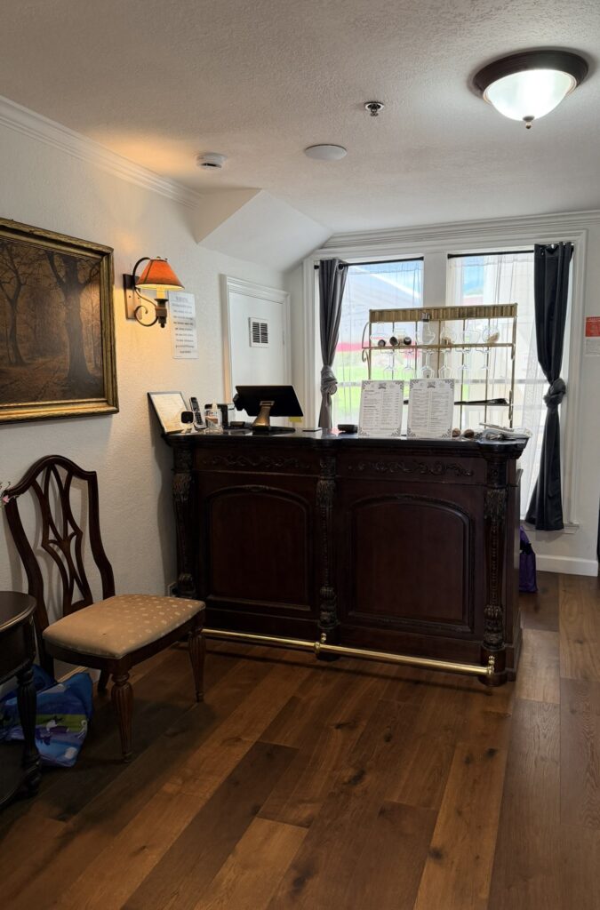 Mountain View Inn reception area with a dark wooden desk holding menus, a computer, and a chair nearby, framed artwork on the wall, and windows with curtains. Warm lighting illuminates the space.