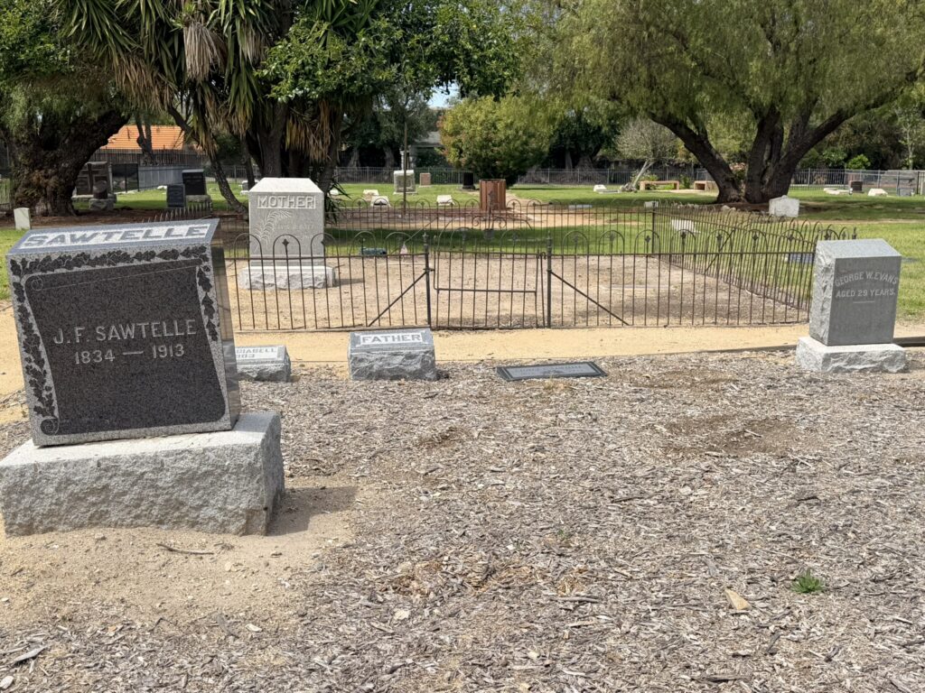 Gravestones for J.F. Sawtelle (1834-1913), Mother, Father, and George W. Evans (aged 29 years) in a fenced cemetery plot with trees, part of a pioneer family memorial.
