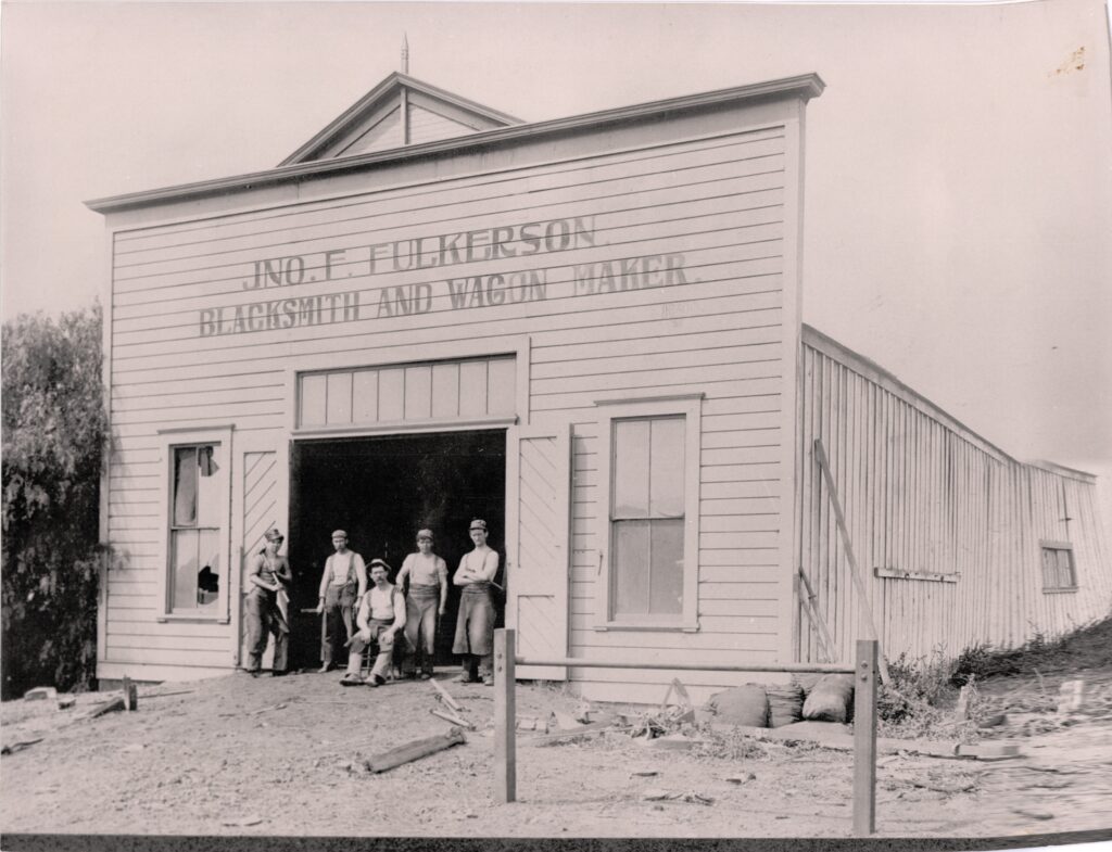 1902 black-and-white photograph of Jno. F. Fulkerson's Somis Blacksmith Shop, showing five men standing in front of the open garage door with "BLACKSMITH AND WAGON MAKER" lettering on white siding.