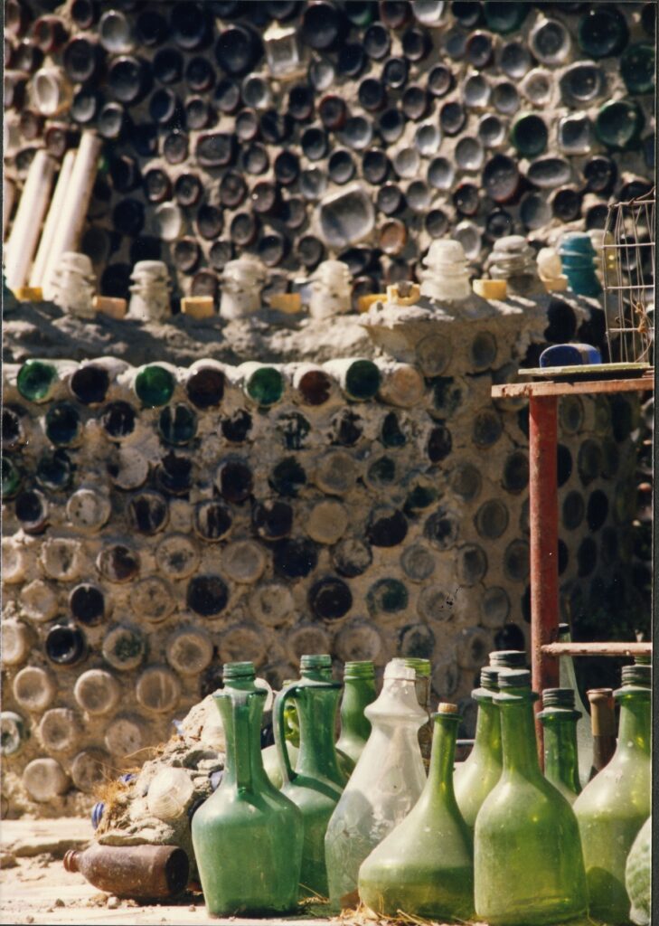 A mosaic wall constructed from assorted glass bottles and jars in green, brown, and clear hues, with additional green glass bottles and jars arranged on a sandy surface in the foreground; detail photo from circa 1980s.