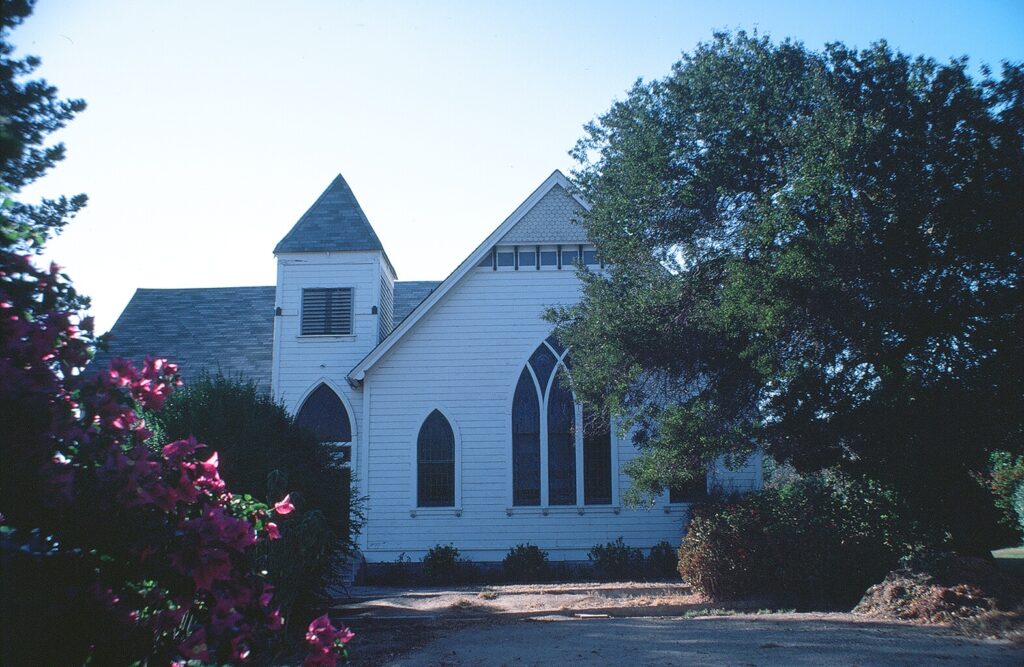 White wooden church with Gothic windows and steeple, surrounded by trees and pink flowers, photographed in 1981.