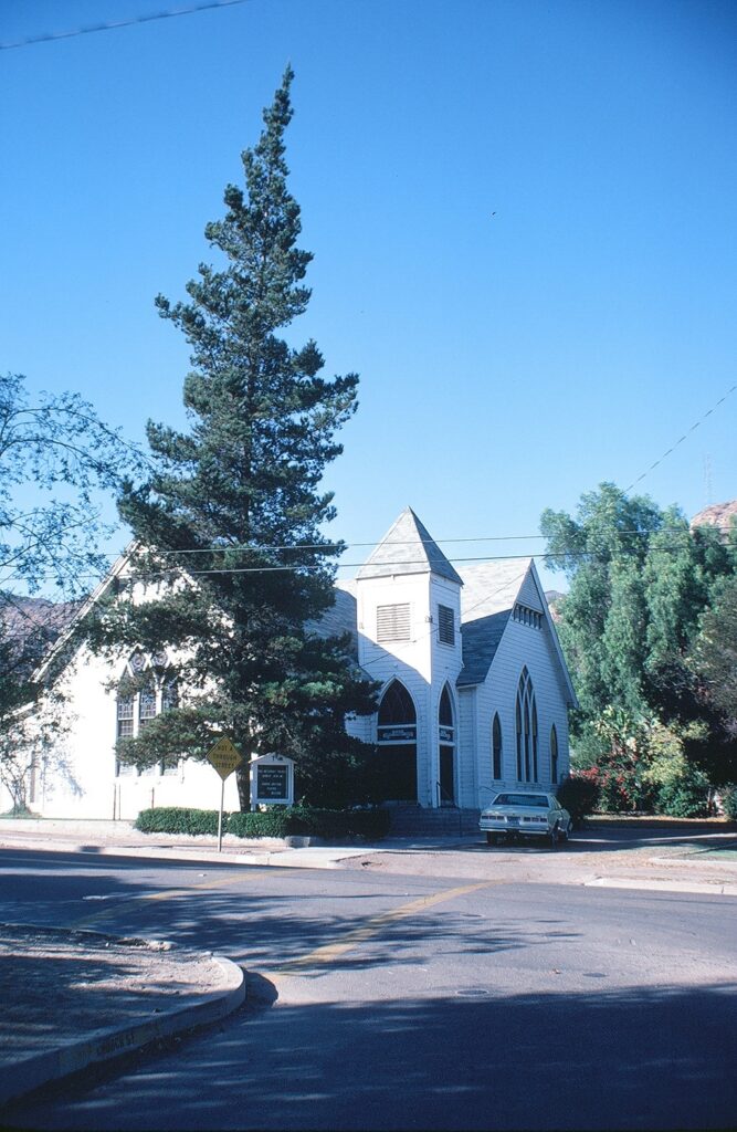 A 1981 photograph of a white church with a steeple and arched windows, partially obscured by a tall pine tree. A "No Through Street" sign and a parked car are visible on the street corner under a clear blue sky.