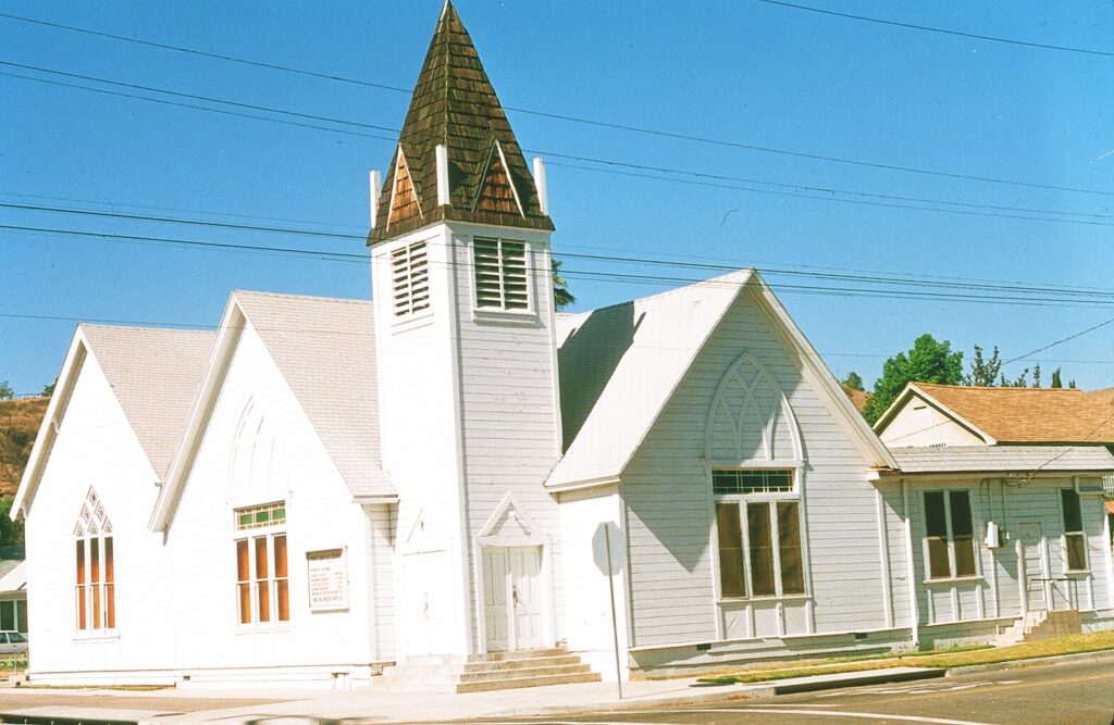 White Methodist church building from 1985 with a steeple, Gothic windows, and a signboard on a street with power lines under a clear blue sky.