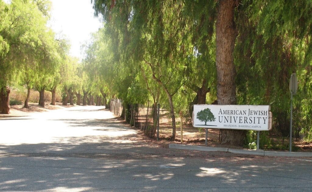 A sunlit tree-lined road with a white sign for American Jewish University's Brandeis-Bardin Campus, featuring a green tree icon and black text.