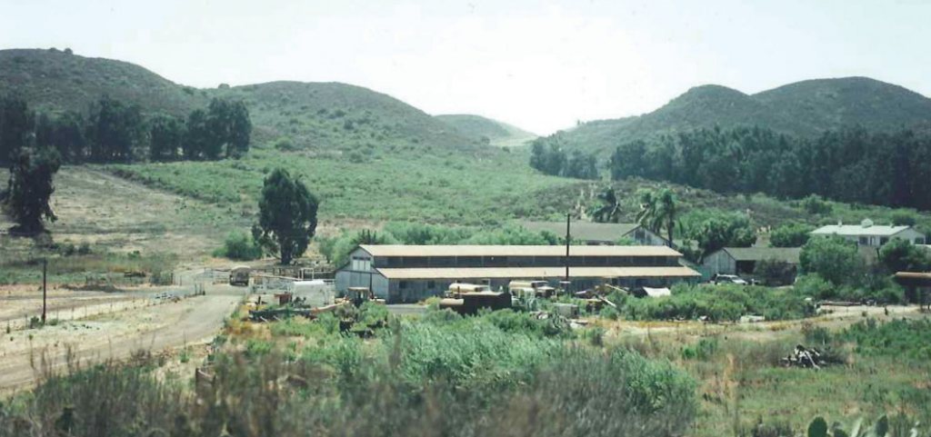 Dos Vientos Ranch circa 1980s features a large building complex with a dirt road, surrounded by hills, trees, and scattered structures in a rural landscape.