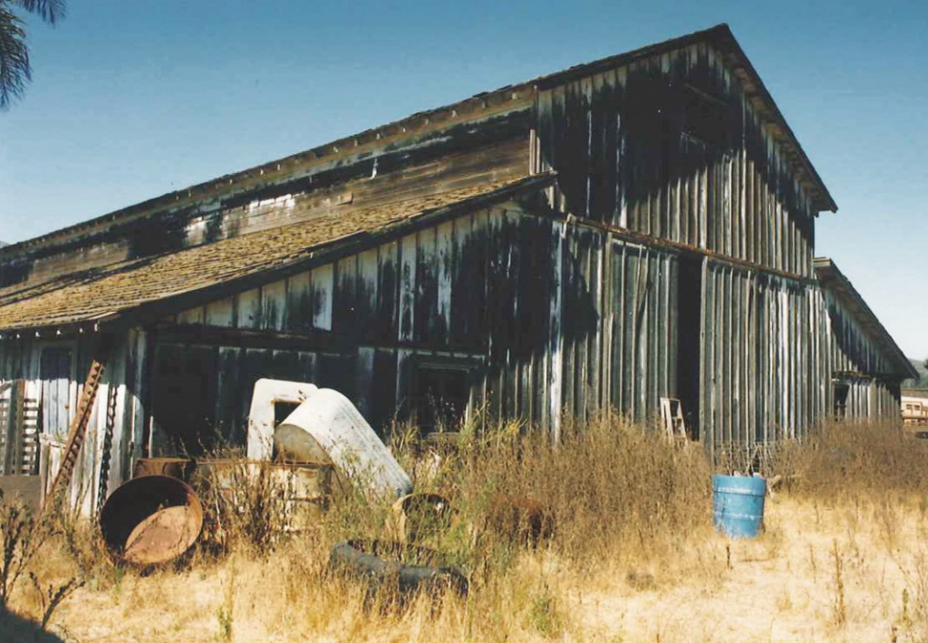 A weathered barn with rusted metal siding, overgrown dry grass, and scattered farm equipment including barrels and a blue container, part of the Dos Vientos Ranch circa 1980s.