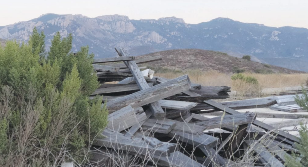 A pile of weathered wooden beams and planks, remnants of the Dos Vientos Ranch Buildings, lies in Rancho Portrero open space with surrounding vegetation and mountains in the background.