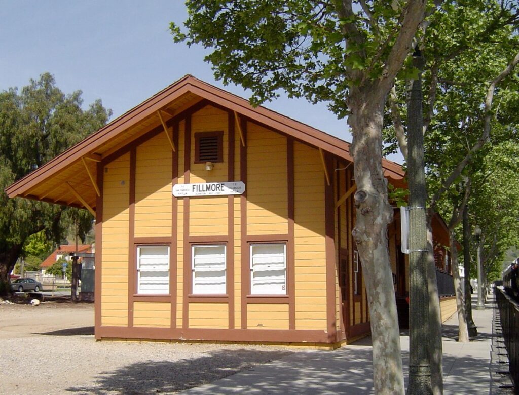 Yellow and brown wooden railroad depot with a "FILLMORE" sign, three windows with white shutters, and a covered porch, situated along railroad tracks with trees in the foreground.
