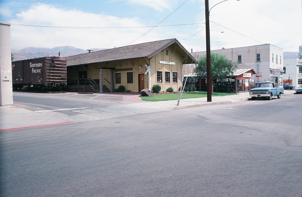 Yellow Fillmore Southern Pacific Railroad Depot with a brown "Southern Pacific" freight car on the left, a railroad crossing sign, and a blue pickup truck on the street, relocated in the 1970s.