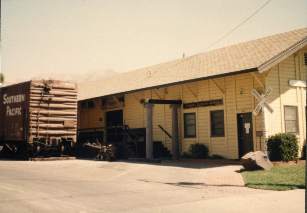 Yellow Southern Pacific Railroad Depot building with "Railway Express Agency" signage, a Southern Pacific freight car to the left, and a railroad crossing sign to the right, relocated circa 1980s.