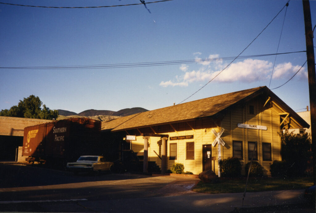 A yellow Southern Pacific Railroad Depot building with "Railway Express Agency" signage, a "FILLMORE" street sign, and a red freight car, relocated circa 1980s with mountains and power lines in the background.