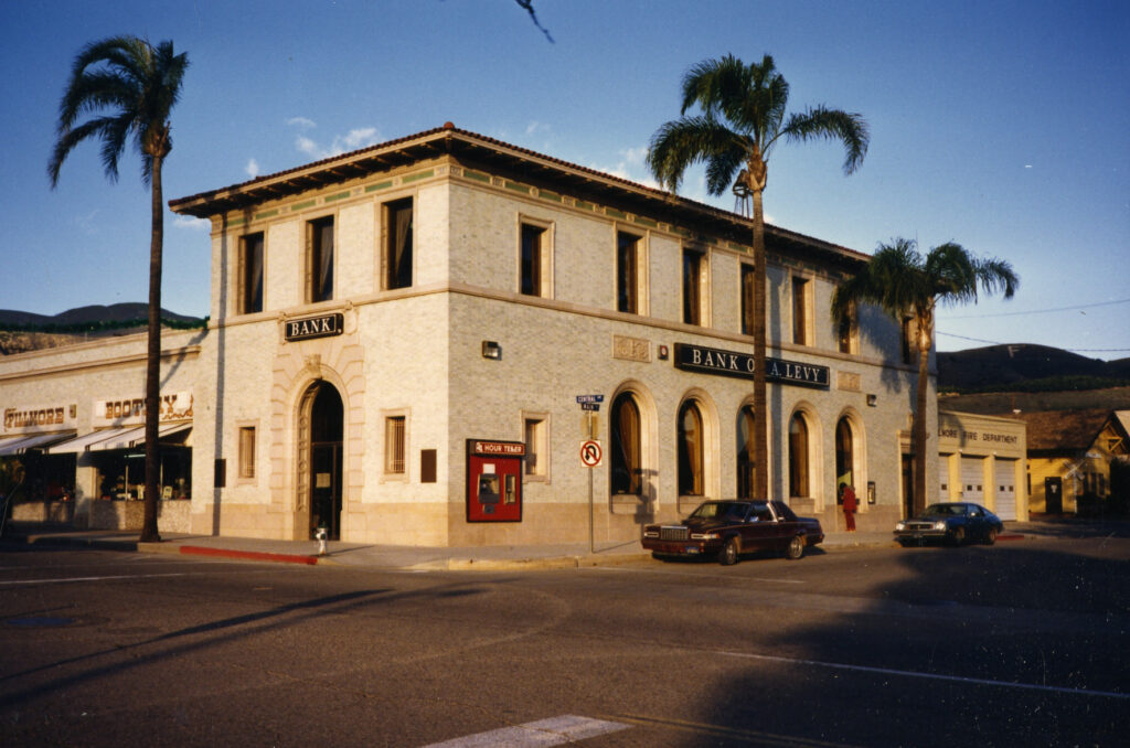 Two-story Fillmore State Bank building from 1986, featuring "BANK" and "BANK OF A. LEVY" signs, a red ATM, palm trees, parked cars, and adjacent storefronts including "FILLMORE" and "BOOT".