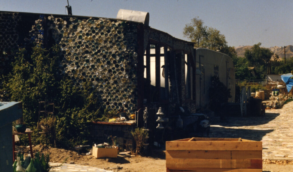 A house with walls made of recycled glass bottles, a trailer, and garden items in Grandma Prisbrey's Bottle Village from the 1980s.