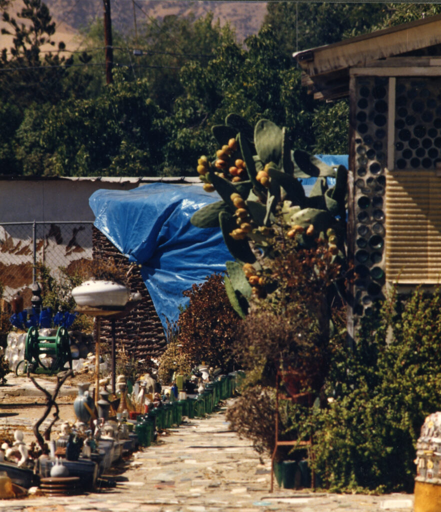 A stone path winds through Grandma Prisbrey's 1980s bottle village, lined with bottle sculptures, cacti bearing yellow fruit, and a blue tarp-covered structure.