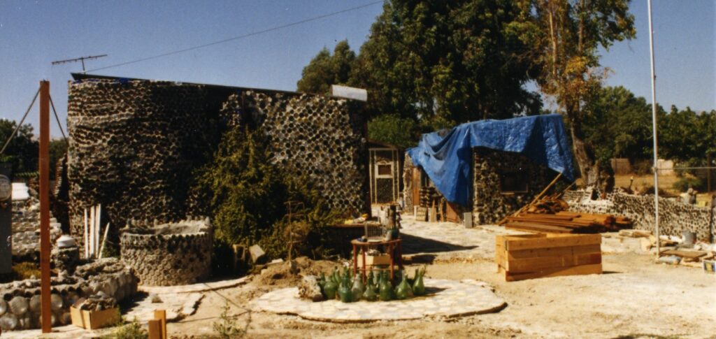Construction site of Grandma Prisbrey's Bottle Village (1980s) features bottle masonry walls, a blue tarp-covered structure, green glass bottles on a circular patio, and wooden planks under a clear sky.