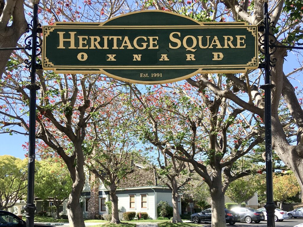 Green sign with gold text "Heritage Square Oxnard Est. 1991" on black posts, with trees and houses in the background.
