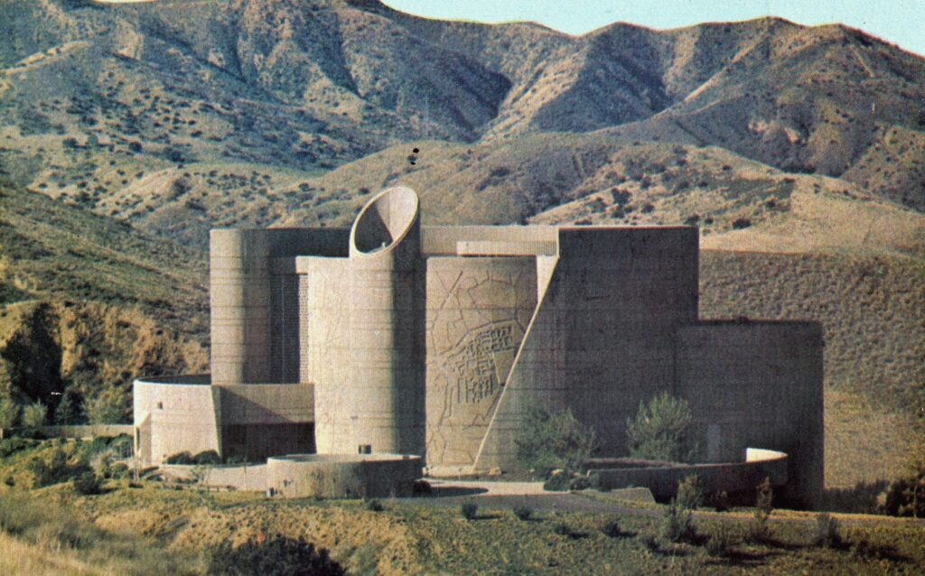 Modernist concrete building, the House of the Book (1978), features a cylindrical tower with a circular opening and etched designs on a large wall, set against a mountainous landscape.