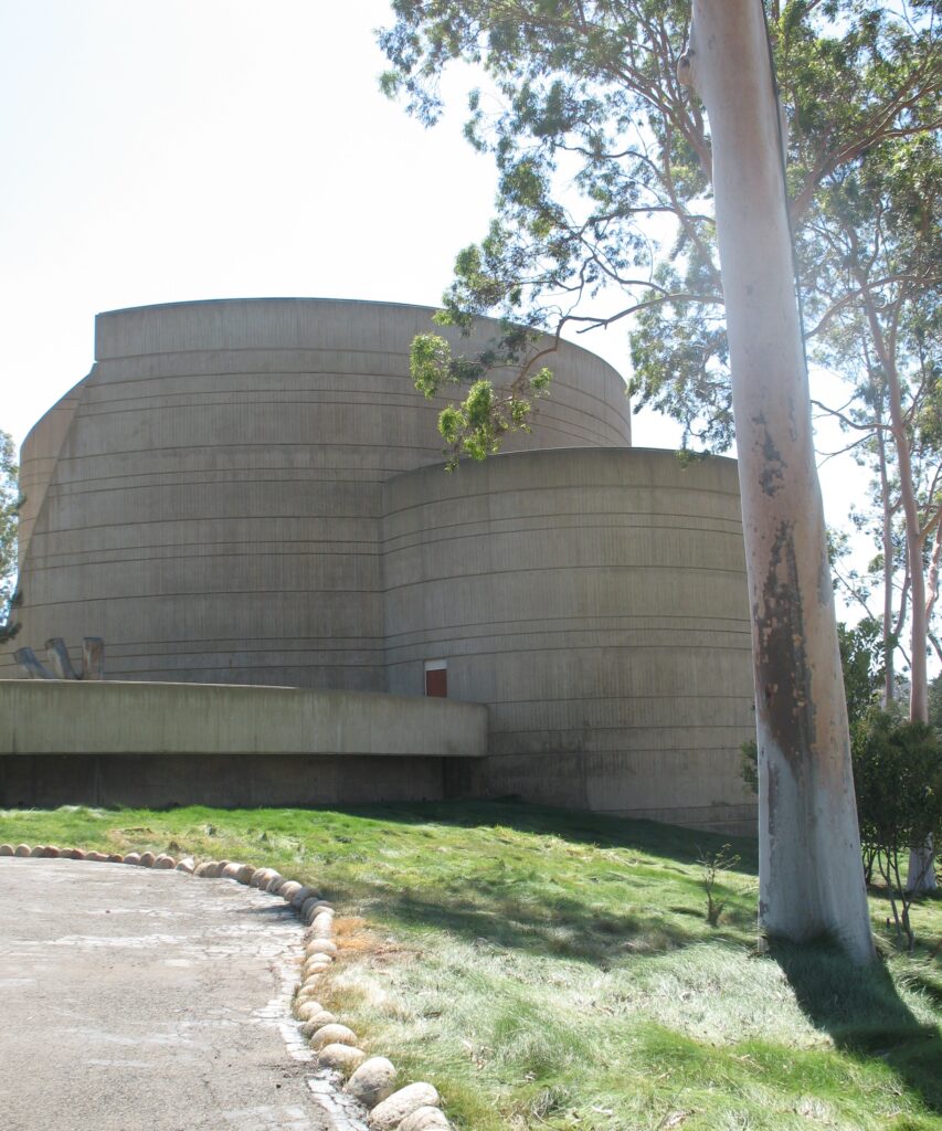 A large curved concrete building with horizontal lines and a small red door, situated on a grassy slope with a paved path edged by stones and trees in the foreground.