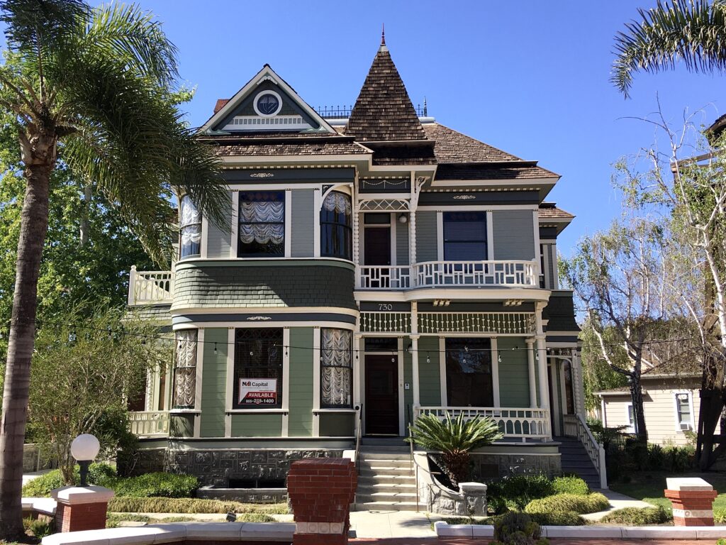 A green Victorian-style house with white trim, a turret, balconies, and address '730' displays a 'Mai Capital AVAILABLE' sign.