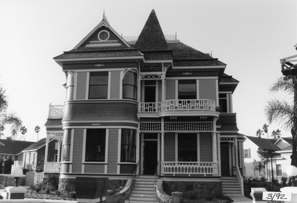 Black-and-white photograph of the Justin Petit Ranch House at Heritage Square, 1992, featuring Victorian architecture with wraparound balconies, a turret, and palm trees in the background; '3/92' date stamp visible in bottom right corner.